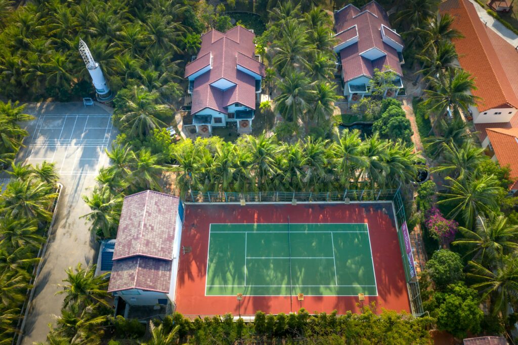 Discover a vibrant tennis court nestled amid lush palm trees and tropical residences from a stunning aerial perspective.